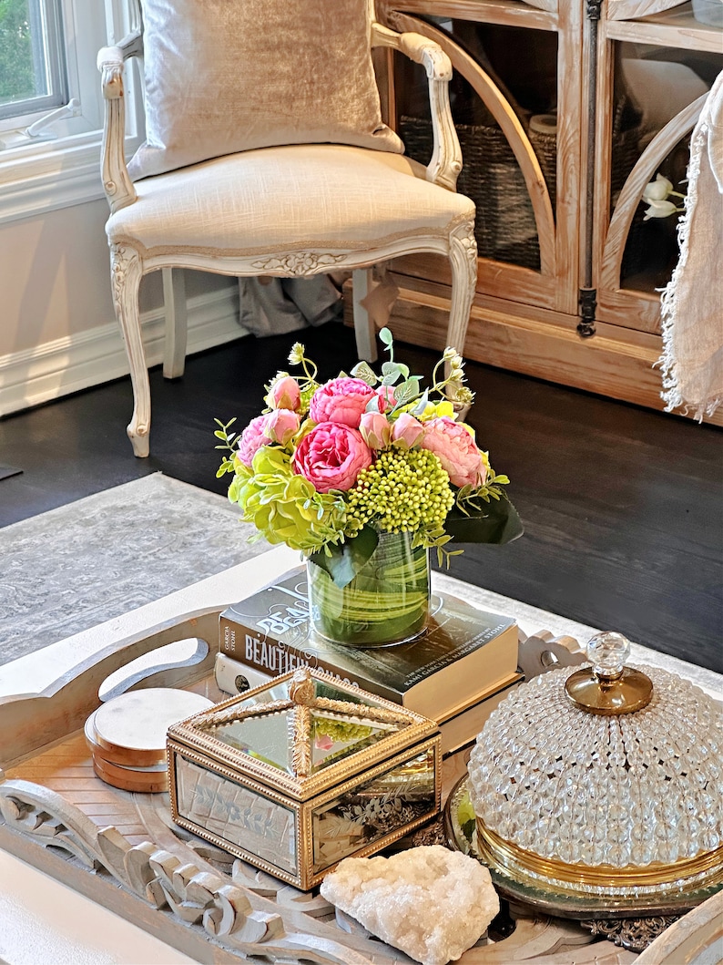 Op de afbeelding: Een houten dienblad met een decoratief ontwerp heeft een kristallen koepel, een spiegeldoos, een ronde houten onderzetter en een vaas met roze en groene bloemen. Het dienblad staat op een salontafel met een boek en een witte steen.