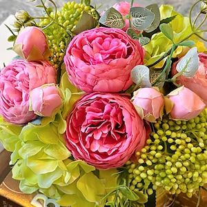 Op de afbeelding: Een ronde bloemstuk met roze pioenen en groene hortensia's. Het bloemstuk staat op een boek met het zichtbare woord "BEAUTIFUL".