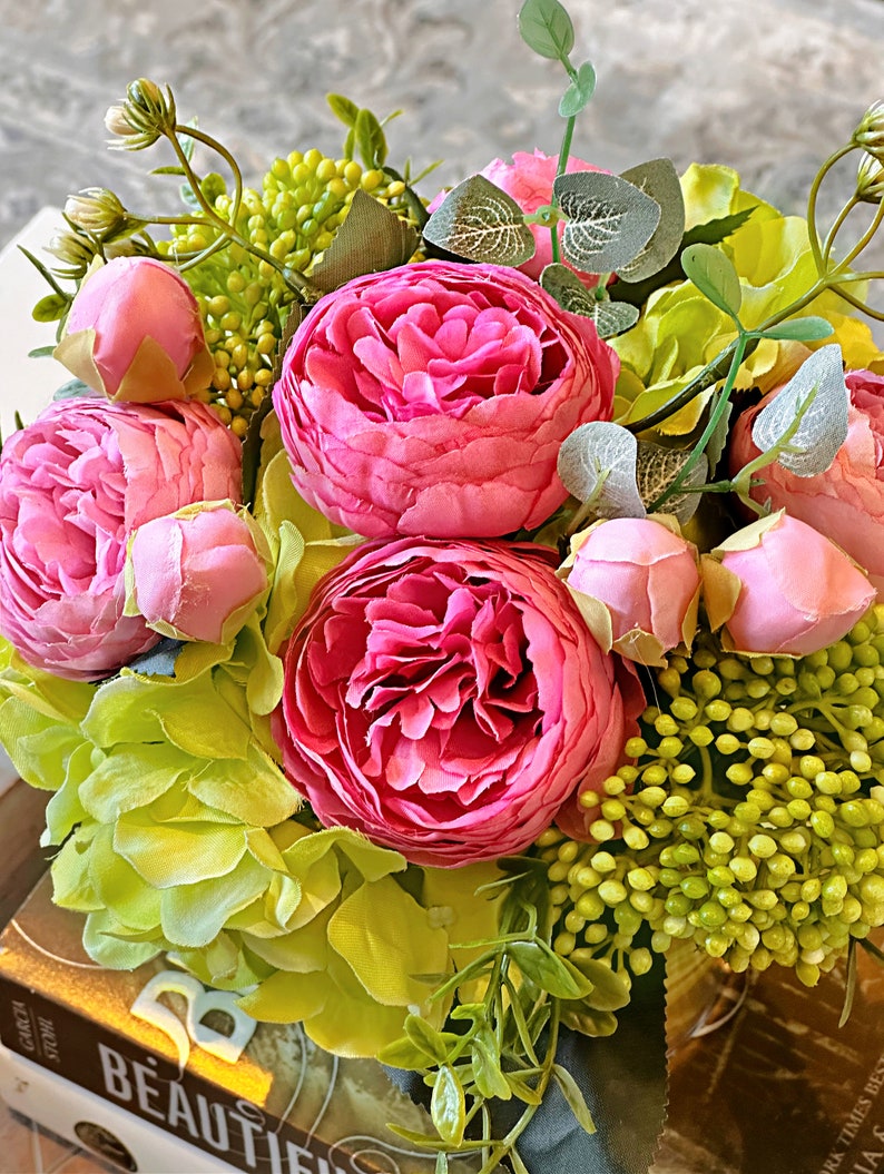 Op de afbeelding: Een ronde bloemstuk met roze pioenen en groene hortensia's. Het bloemstuk staat op een boek met het zichtbare woord "BEAUTIFUL".
