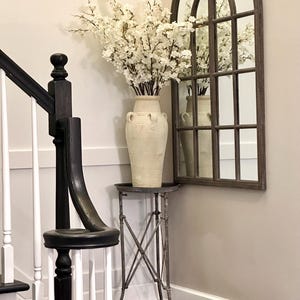 May include: A white ceramic vase with a floral arrangement of white blossoms sits on a small metal table with an arched mirror in the background.