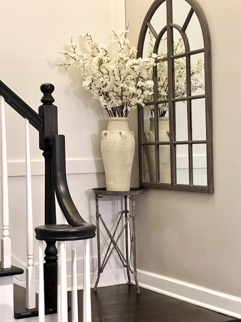 May include: A white ceramic vase with a floral arrangement of white blossoms sits on a small metal table with an arched mirror in the background.