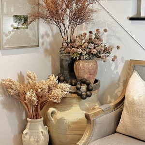 May include: A decorative arrangement featuring various vases and dried floral elements. The display includes a large, light-colored ceramic jar, smaller vases in shades of gray and brown, and a gray upholstered chair. The arrangement is placed near a staircase.