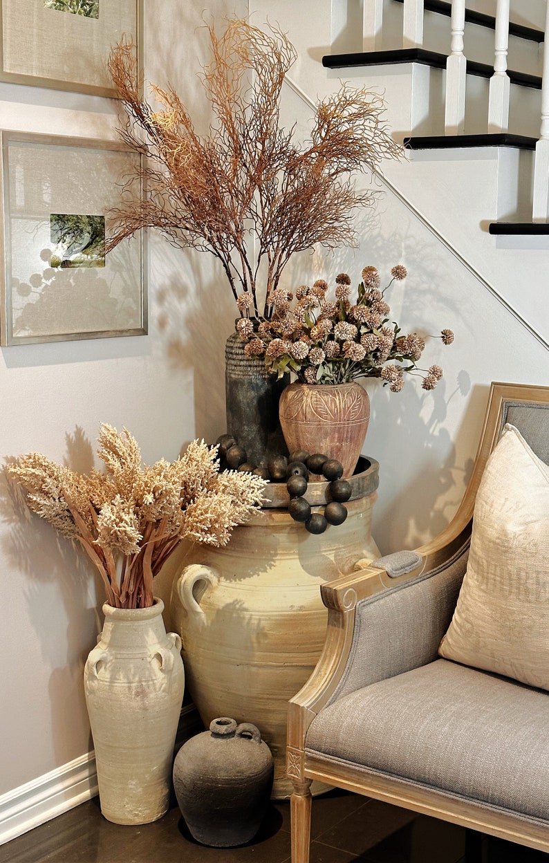 May include: A decorative arrangement featuring various vases and dried floral elements. The display includes a large, light-colored ceramic jar, smaller vases in shades of gray and brown, and a gray upholstered chair. The arrangement is placed near a staircase.