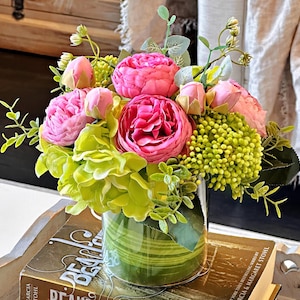 Op de afbeelding: Een kunstbloemenarrangement in een glazen vaas met een groen gestreept patroon. De vaas staat op een boek met het woord "Deals" zichtbaar. De bloemen zijn roze en groen.
