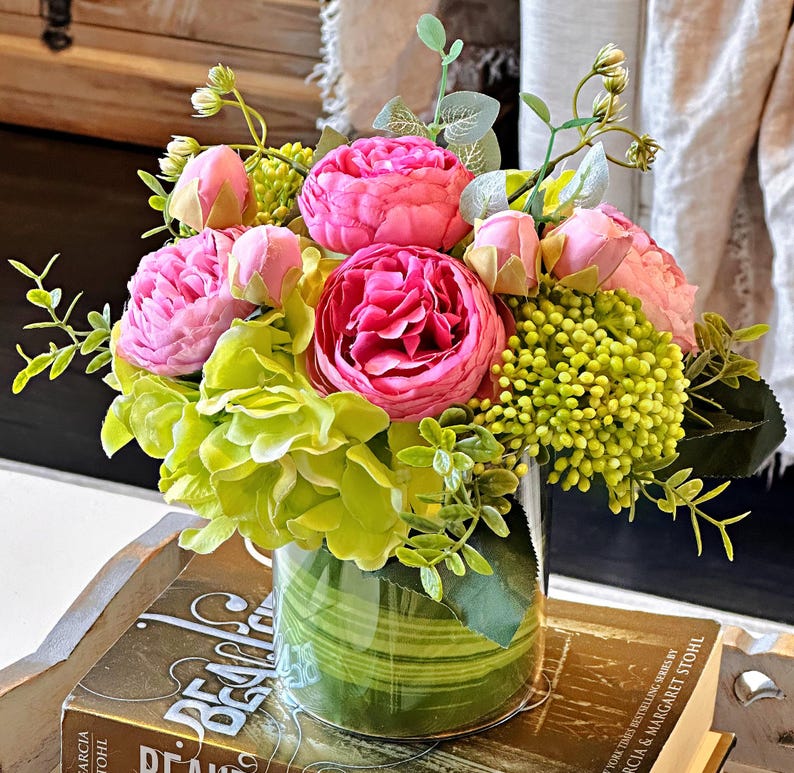 Op de afbeelding: Een kunstbloemenarrangement in een glazen vaas met een groen gestreept patroon. De vaas staat op een boek met het woord "Deals" zichtbaar. De bloemen zijn roze en groen.