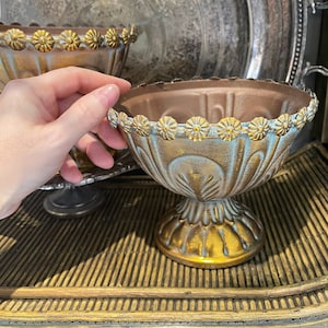 May include: Two decorative metal bowls with a distressed gold and blue finish. The bowls have a scalloped edge with a row of gold flower accents. The bowls are on a wooden tray with a woven pattern.