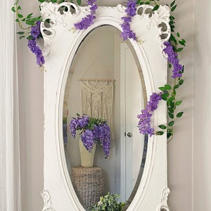Op de afbeelding: Een sierlijke, witte ovale spiegel is versierd met paarse wisteria slingers en groene bladeren. De spiegel weerspiegelt een kamer met een macram&eacute; wandkleed en een vaas met paarse bloemen. De tekst "One garland" staat linksboven.