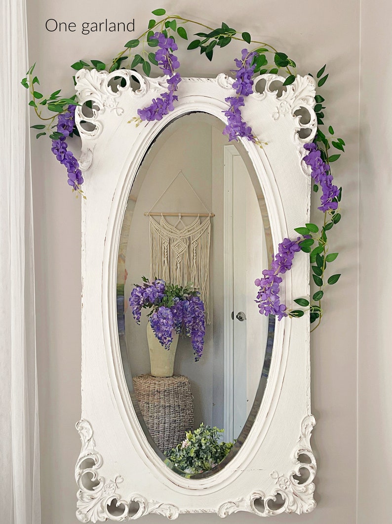 Op de afbeelding: Een sierlijke, witte ovale spiegel is versierd met paarse wisteria slingers en groene bladeren. De spiegel weerspiegelt een kamer met een macram&eacute; wandkleed en een vaas met paarse bloemen. De tekst "One garland" staat linksboven.