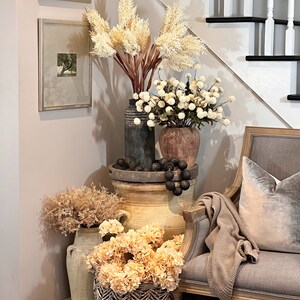 May include: A decorative corner arrangement featuring various vases and baskets filled with dried flowers and faux blooms in neutral tones. A gray upholstered chair with a velvet pillow and a knitted throw is positioned nearby. Two framed pictures hang on the wall.