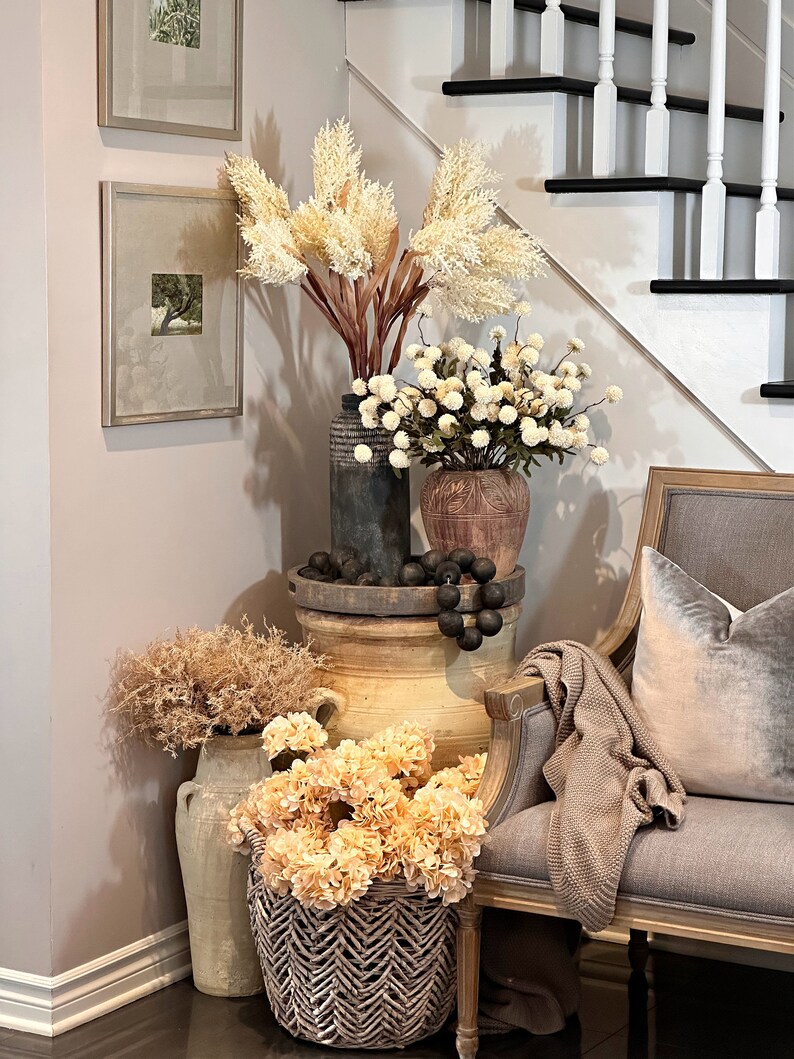 May include: A decorative corner arrangement featuring various vases and baskets filled with dried flowers and faux blooms in neutral tones. A gray upholstered chair with a velvet pillow and a knitted throw is positioned nearby. Two framed pictures hang on the wall.