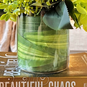 Op de afbeelding: Een heldere glazen vaas met een groen-wit gestreept lint eromheen gewikkeld. De vaas is gevuld met een boeket bloemen, waaronder roze rozen, groene hortensia's en groen blad. De vaas staat op een boek met de titel "Beautiful Chaos".