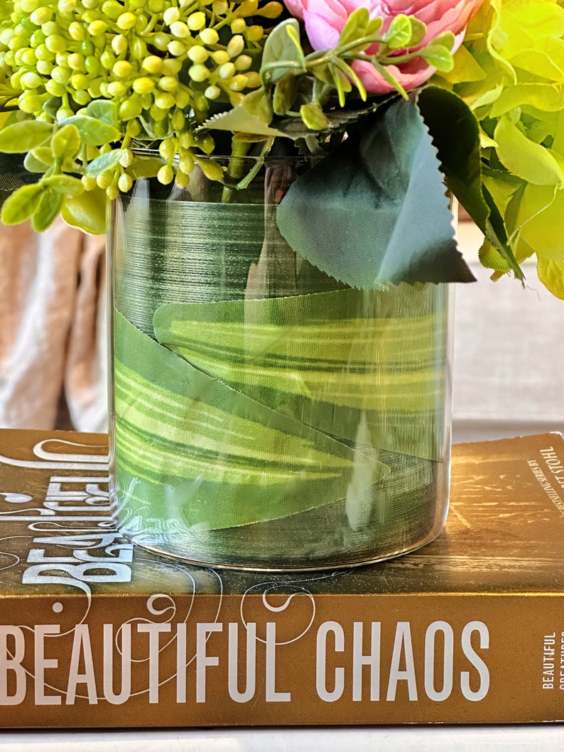 Op de afbeelding: Een heldere glazen vaas met een groen-wit gestreept lint eromheen gewikkeld. De vaas is gevuld met een boeket bloemen, waaronder roze rozen, groene hortensia's en groen blad. De vaas staat op een boek met de titel "Beautiful Chaos".