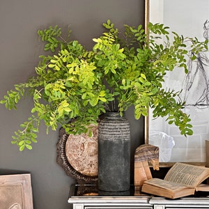 May include: A dark gray ceramic vase with a textured surface holds a bouquet of green leafy branches. The vase is sitting on a silver mirrored dresser with a framed print and a book on top.