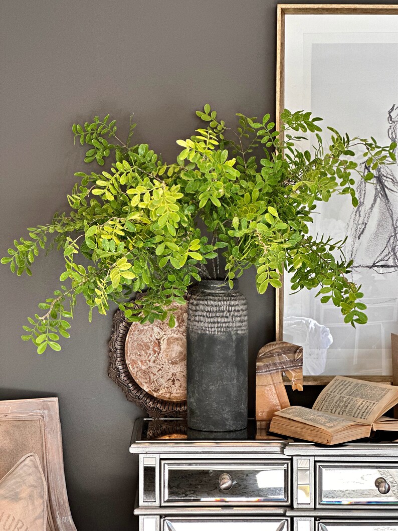 May include: A dark gray ceramic vase with a textured surface holds a bouquet of green leafy branches. The vase is sitting on a silver mirrored dresser with a framed print and a book on top.