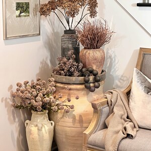May include: A decorative corner arrangement featuring a large, aged terracotta vase filled with dried flowers. Other vases of varying sizes and textures hold additional dried floral arrangements. A framed picture and a bench with a throw blanket complete the scene.