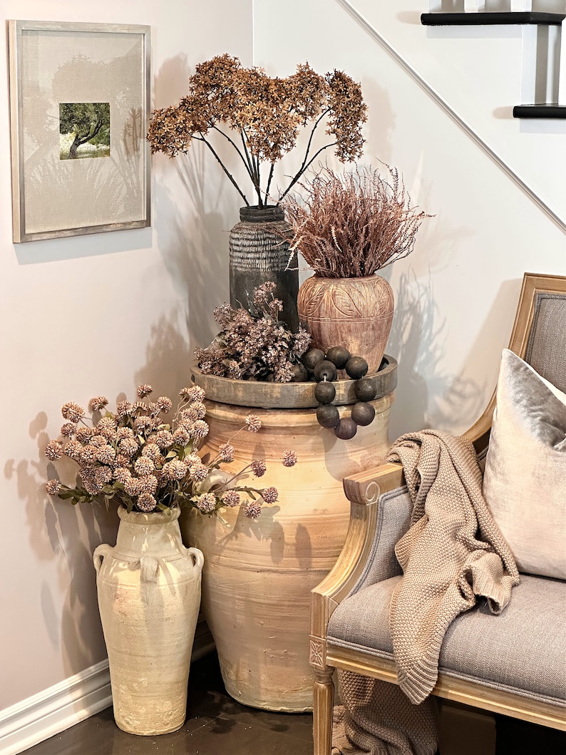 May include: A decorative corner arrangement featuring a large, aged terracotta vase filled with dried flowers. Other vases of varying sizes and textures hold additional dried floral arrangements. A framed picture and a bench with a throw blanket complete the scene.