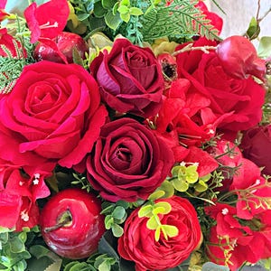 May include: A close-up of a floral arrangement dominated by deep red roses and smaller red flowers, with green foliage. The arrangement includes red apples, adding to the rich colour palette. The flowers are densely packed, creating a full and lush appearance.
