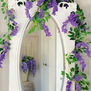 Op de afbeelding: Een ovale spiegel met een wit frame, versierd met paarse wisteria-ranken en groene bladeren. De spiegel weerspiegelt een kamer met een macram&eacute; wandkleed en een vaas met wisteria. De spiegel heeft een vintage uitstraling.
