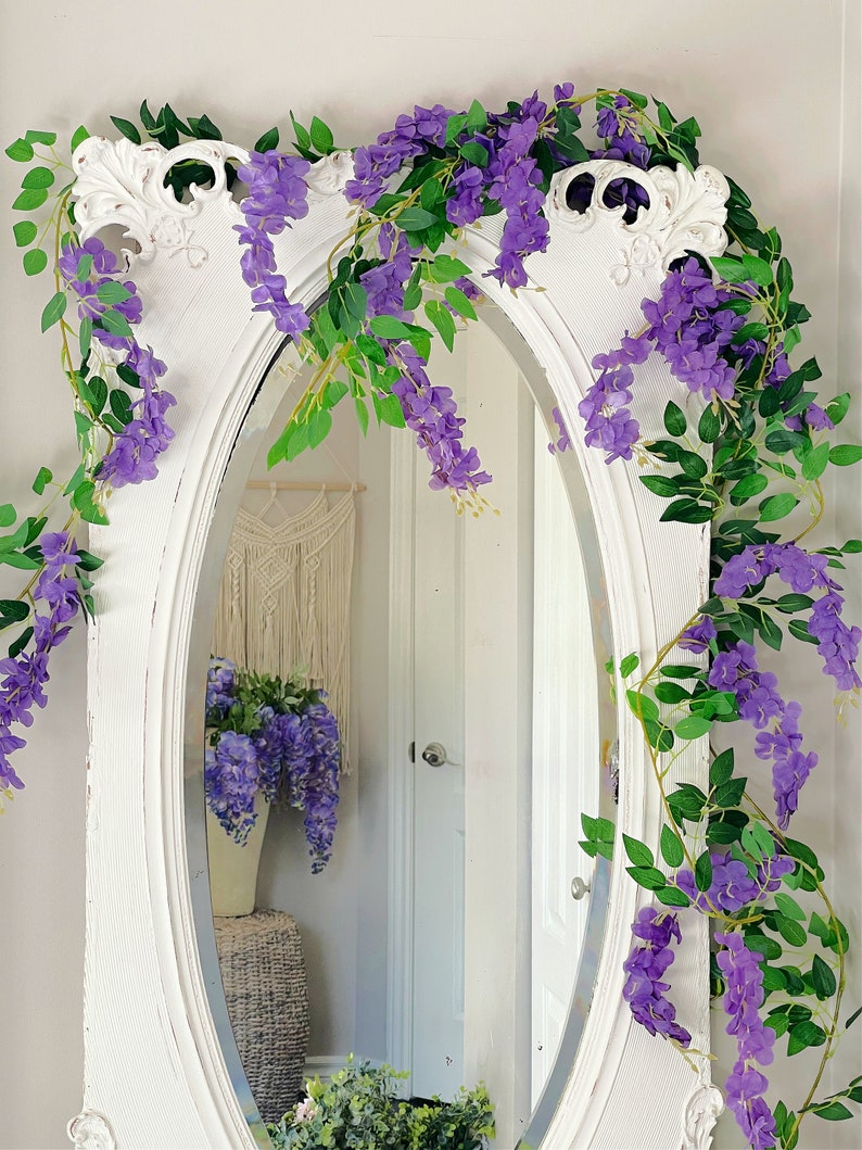 Op de afbeelding: Een ovale spiegel met een wit frame, versierd met paarse wisteria-ranken en groene bladeren. De spiegel weerspiegelt een kamer met een macram&eacute; wandkleed en een vaas met wisteria. De spiegel heeft een vintage uitstraling.