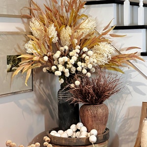 May include: A decorative arrangement featuring dried flowers and faux foliage in various vases. The display includes a large black vase, a brown carved vase, and a wooden tray with white orbs. The color palette includes cream, brown, and gold tones.