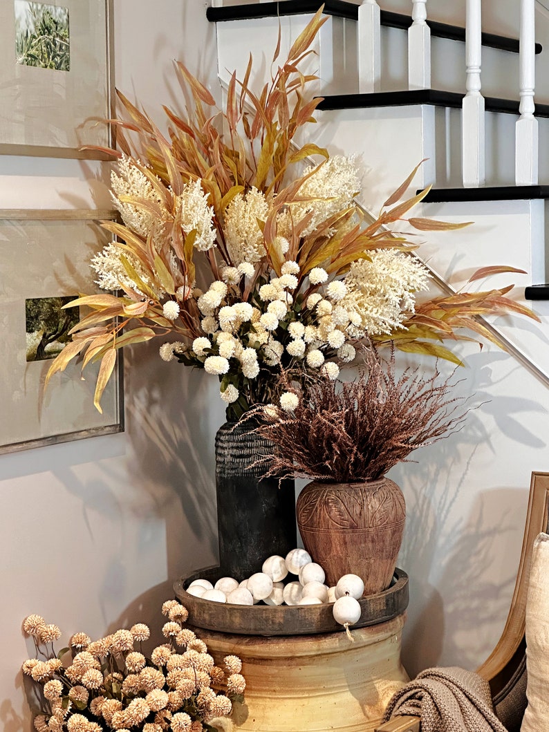 May include: A decorative arrangement featuring dried flowers and faux foliage in various vases. The display includes a large black vase, a brown carved vase, and a wooden tray with white orbs. The color palette includes cream, brown, and gold tones.