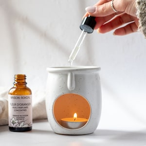 May include: A white ceramic oil diffuser with a small candle inside. A brown glass bottle of orange blossom scented oil with a dropper is being poured into the diffuser.