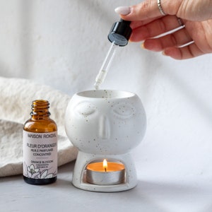 Puede incluir: Difusor de cerámica blanca con un diseño de cara y una vela pequeña en el interior. Una botella de vidrio marrón de aceite perfumado de flor de azahar con un gotero se está utilizando para agregar aceite al difusor.