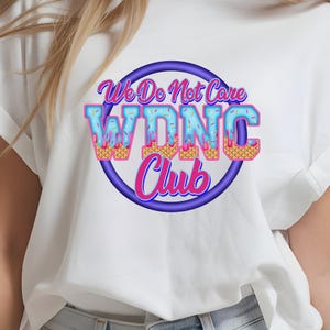 May include: White t-shirt featuring a circular graphic with the text "We Do Not Care WDNC Club." The text is in a playful font with blue, pink, and yellow accents, resembling ice cream. The graphic is outlined in purple.