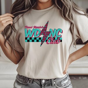 May include: Beige t-shirt with the text "Proud Member WDNC Club". The design features a teal and pink lightning bolt with a leopard print pattern, and teal and black checkered stripes.