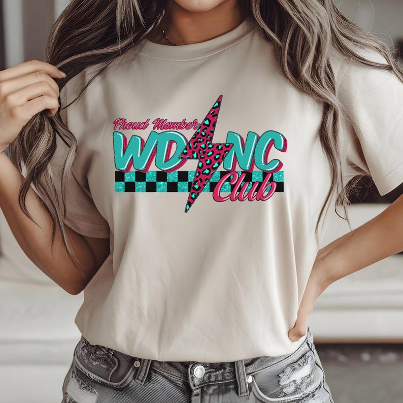 May include: Beige t-shirt with the text "Proud Member WDNC Club". The design features a teal and pink lightning bolt with a leopard print pattern, and teal and black checkered stripes.