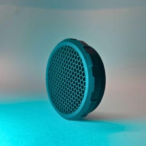May include: A black honeycomb grid lens attachment for a camera flash. The grid is made of plastic and has a hexagonal pattern.