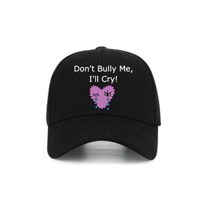 Könnte beinhalten: Schwarze Baseballkappe mit dem Text "Don't Bully Me, I'll Cry!" und einem rosa Herz mit einem blauen Auge und blauen Tränen.