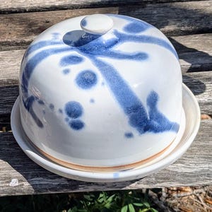 Peut inclure: Un beurrier en céramique blanche avec un couvercle en forme de dôme et un petit bouton. Le plat et le couvercle sont décorés de coups de pinceau et de taches bleues. Le plat repose sur une base ronde.