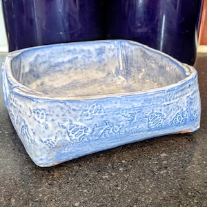Op de afbeelding: Een lichtblauwe keramische bakvorm met een reliëf van bladeren en bloemen. De vorm is vierkant en heeft een licht afgeronde bodem.