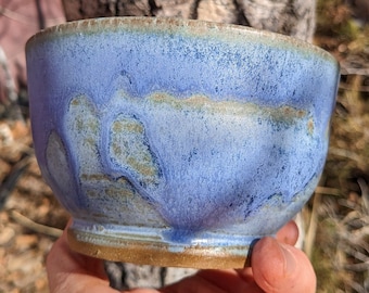 May include: A blue ceramic bowl with a textured glaze. The bowl has a light blue base with darker blue and brown streaks and spots.