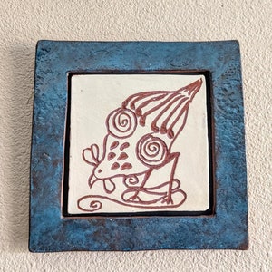 May include: A blue ceramic tile with a white background and a brown line drawing of a stylized bird with a large head and a long tail.