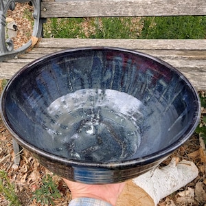 May include: A large, round ceramic bowl featuring a deep, glossy glaze in shades of midnight blue, charcoal, and hints of violet. The interior surface displays a smooth, reflective finish with subtle vertical drip patterns and a dark, solid rim.