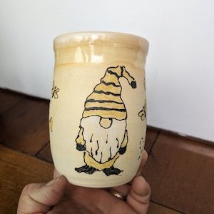 Op de afbeelding: Een keramische mok met een handgeschilderd ontwerp van een gnome met een gele en zwarte gestreepte hoed. De gnome wordt omringd door gele bloemen.