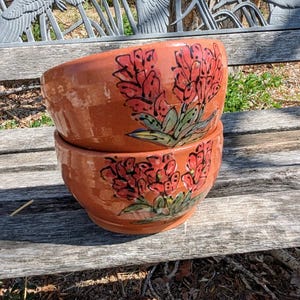 May include: Two terracotta-colored ceramic bowls stacked on top of each other. Each bowl is decorated with a hand-painted floral design in red, green, and blue. The bowls are sitting on a weathered wooden bench.