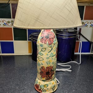 May include: A table lamp with a cream-colored base featuring a hand-painted floral pattern of red flowers, green leaves, and small yellow dots. The lamp is topped with a conical, off-white fabric lampshade with a subtle cross-hatch texture.