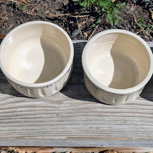May include: Four round ceramic bowls with a cream-colored glaze and a smooth interior. The exterior of each bowl features a vertical ribbed texture and a small, hand-painted blue floral motif consisting of a central dot surrounded by four petals.