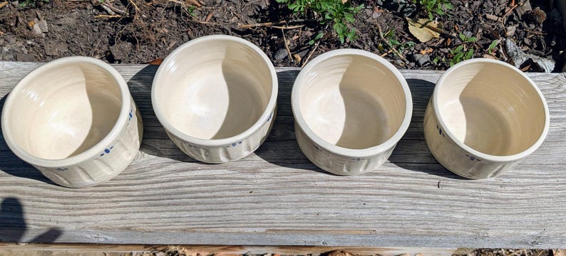 May include: Four round ceramic bowls with a cream-colored glaze and a smooth interior. The exterior of each bowl features a vertical ribbed texture and a small, hand-painted blue floral motif consisting of a central dot surrounded by four petals.