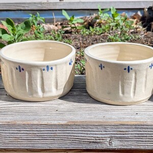 May include: Four cream-colored ceramic bowls arranged in a row. Each bowl features a series of vertical indentations around the exterior and a repeating pattern of small blue painted cross-like marks near the top rim of the vessel.