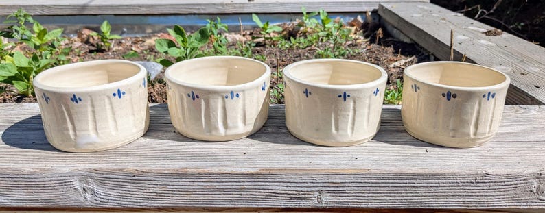 May include: Four cream-colored ceramic bowls arranged in a row. Each bowl features a series of vertical indentations around the exterior and a repeating pattern of small blue painted cross-like marks near the top rim of the vessel.