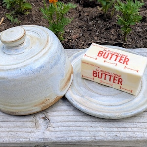 Peut inclure: Un beurrier en céramique fait main, composé d'un couvercle rond et d'une assiette assortie. La poterie présente un émail bleu clair et crème avec de légères nuances brunes. Un bloc rectangulaire de beurre est posé sur l'assiette.