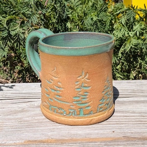 Puede incluir: Una taza de cerámica con cuerpo de arcilla color canela natural y un interior y asa esmaltados en color verde azulado. El exterior presenta siluetas de pinos talladas y rellenas con esmalte verde azulado sobre la forma cilíndrica.