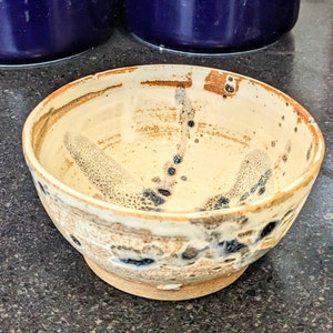 May include: A small, white ceramic bowl with brown and black speckles and a slightly uneven rim.