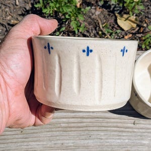 May include: A cream-colored ceramic bowl with a speckled finish, featuring vertical indentations along the exterior wall and a series of small, hand-painted blue cross-shaped marks positioned near the top rim of the vessel.