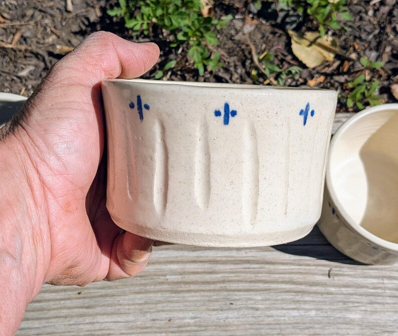 May include: A cream-colored ceramic bowl with a speckled finish, featuring vertical indentations along the exterior wall and a series of small, hand-painted blue cross-shaped marks positioned near the top rim of the vessel.
