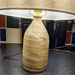 May include: A table lamp featuring a bottle-shaped base with a tan and brown marbled pattern. The lamp includes a gold-toned metal socket and a wide, beige fabric lampshade with a subtle cross-hatch texture, connected by a brown electrical cord.